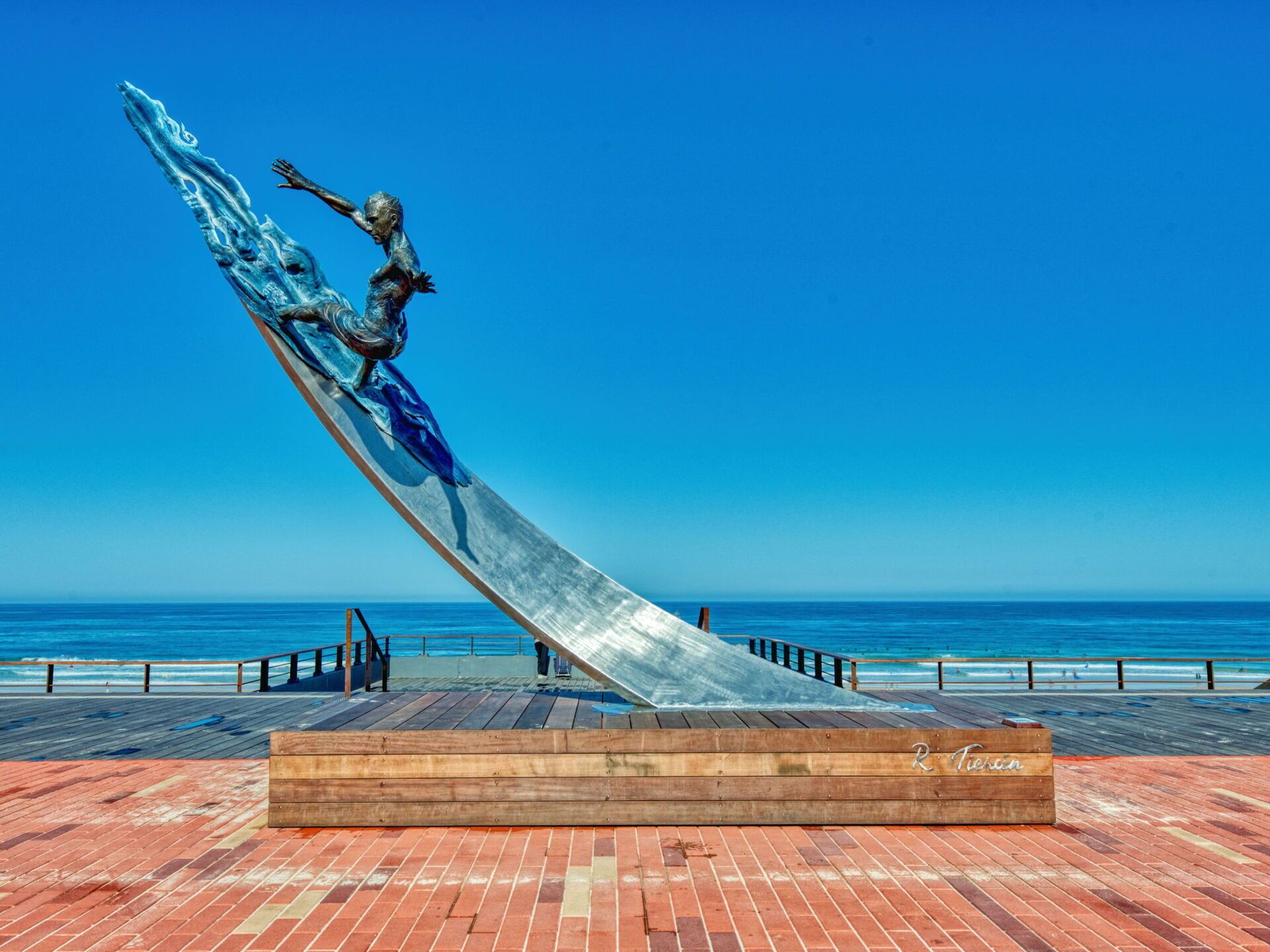 Een sculptuur van een figuur in een dynamische pose op een schuin platform, met een houten bankje en een bord met de tekst 'R. Tiern' op de voorgrond, met uitzicht op een strand.