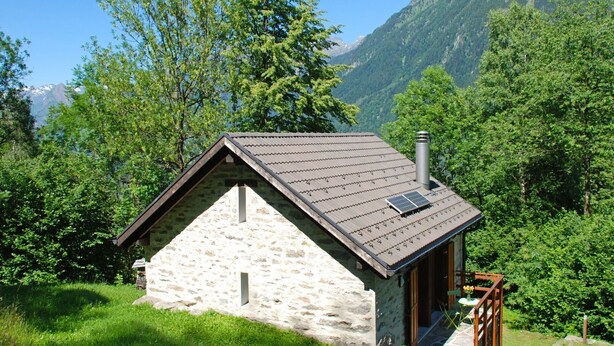 Une petite maison en pierre avec un toit en pente, une cheminée et un petit balcon équipé d’un panneau solaire.