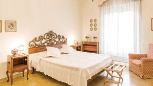 Un dormitorio con una cama grande, una mesita de noche de madera, una silla, un candelabro y una ventana con cortinas.