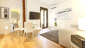 Een moderne woonkamer met een witte bank, een glazen salontafel en een Marshall speaker. Er is een zithoek met witte stoelen en een glazen tafel. Een groot abstract schilderij hangt aan de muur, en een houten zuil is zichtbaar.