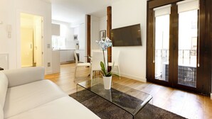 Een moderne woonkamer met een glazen salontafel, een witte bank en een potplant. Er is een eethoek met witte stoelen en een keuken op de achtergrond.