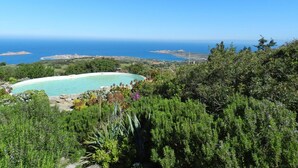 Vista de un paisaje costero con una piscina grande y curvada rodeada de exuberante vegetación y un cielo azul despejado.