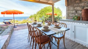 Un espacio de comedor al aire libre con vista al mar, sillas de madera, una mesa con platos y dos sombrillas naranjas.