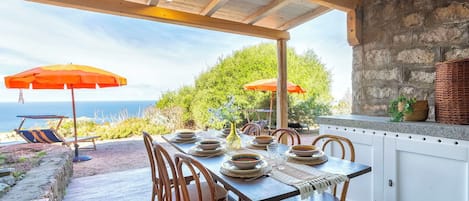 An outdoor dining area with a view of the sea, wooden chairs, a table set with plates, and two orange umbrellas.