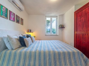 A bedroom with a bed, pillows, a window, a shelf with books, and a red door.