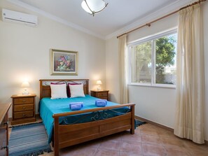 Un dormitorio con una cama de madera, dos mesitas de noche, una lámpara de noche, una ventana con cortinas, una lámpara de techo y un equipo de aire acondicionado.