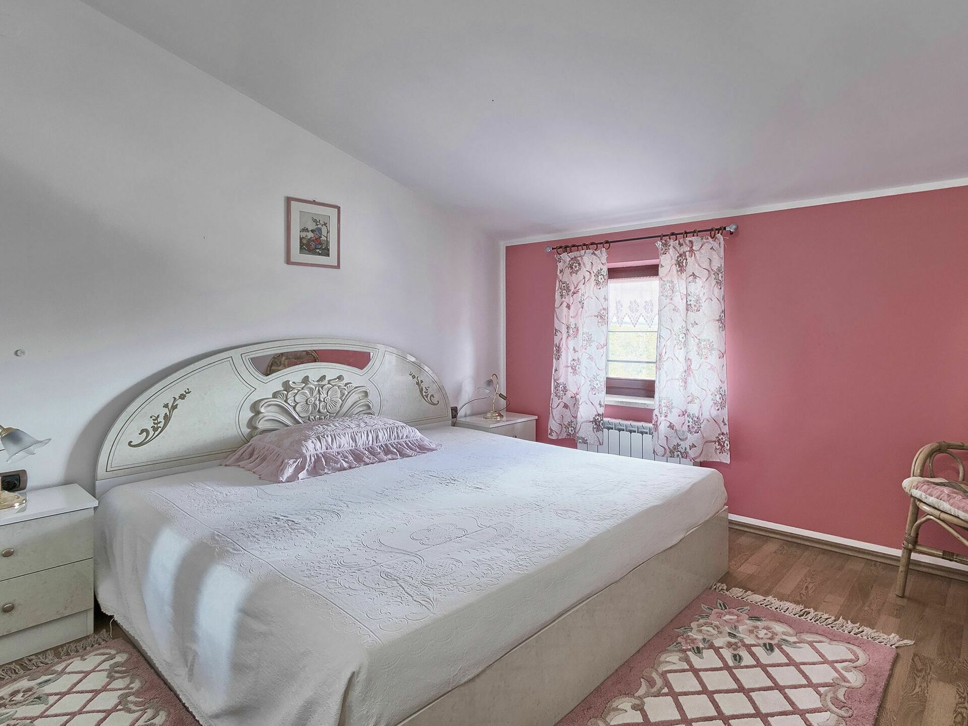 A bedroom with a large bed, a nightstand, a dresser, and a window with floral curtains.
