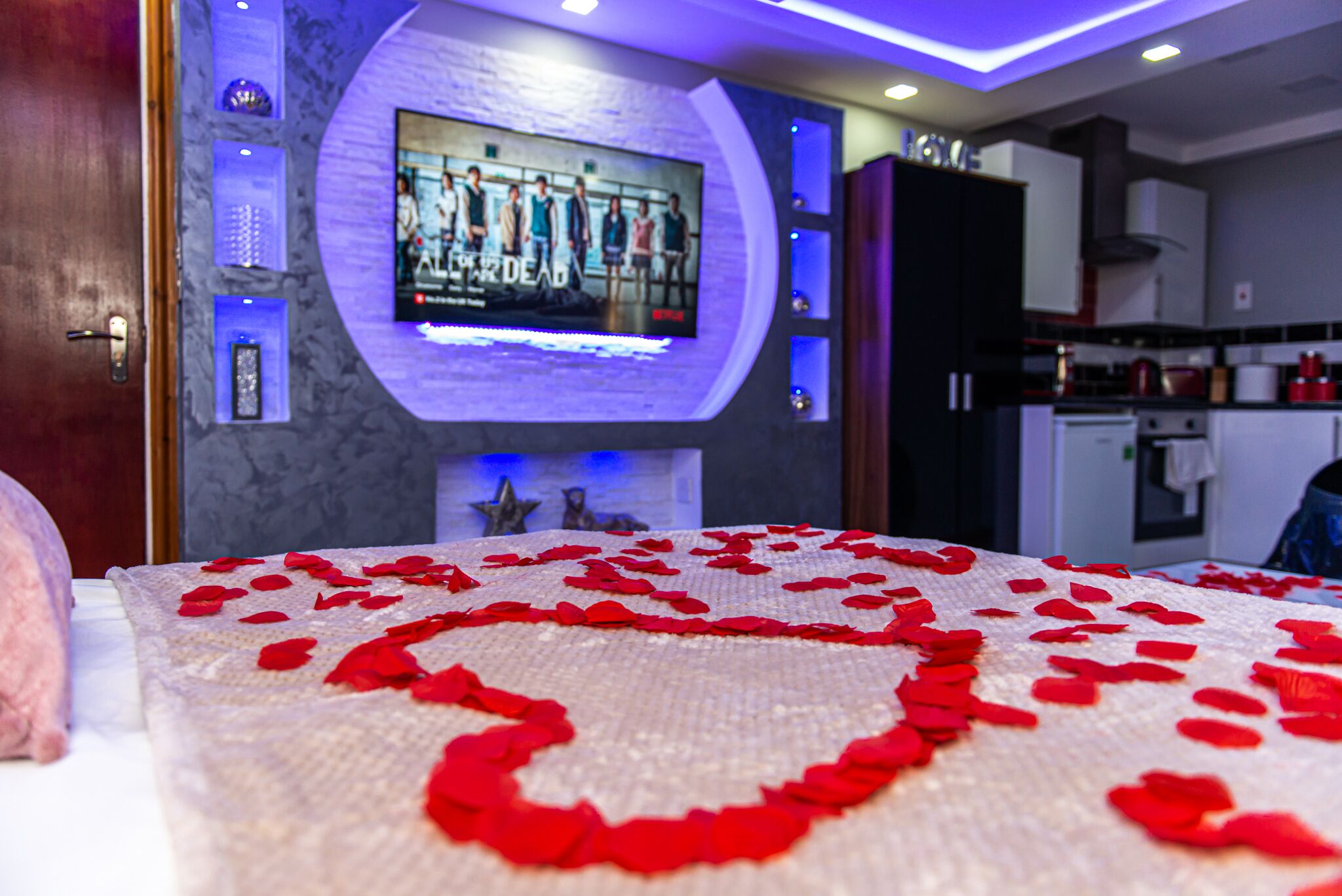 A hotel room with a bed adorned with rose petals, a television mounted on the wall, and a kitchen area visible in the background.