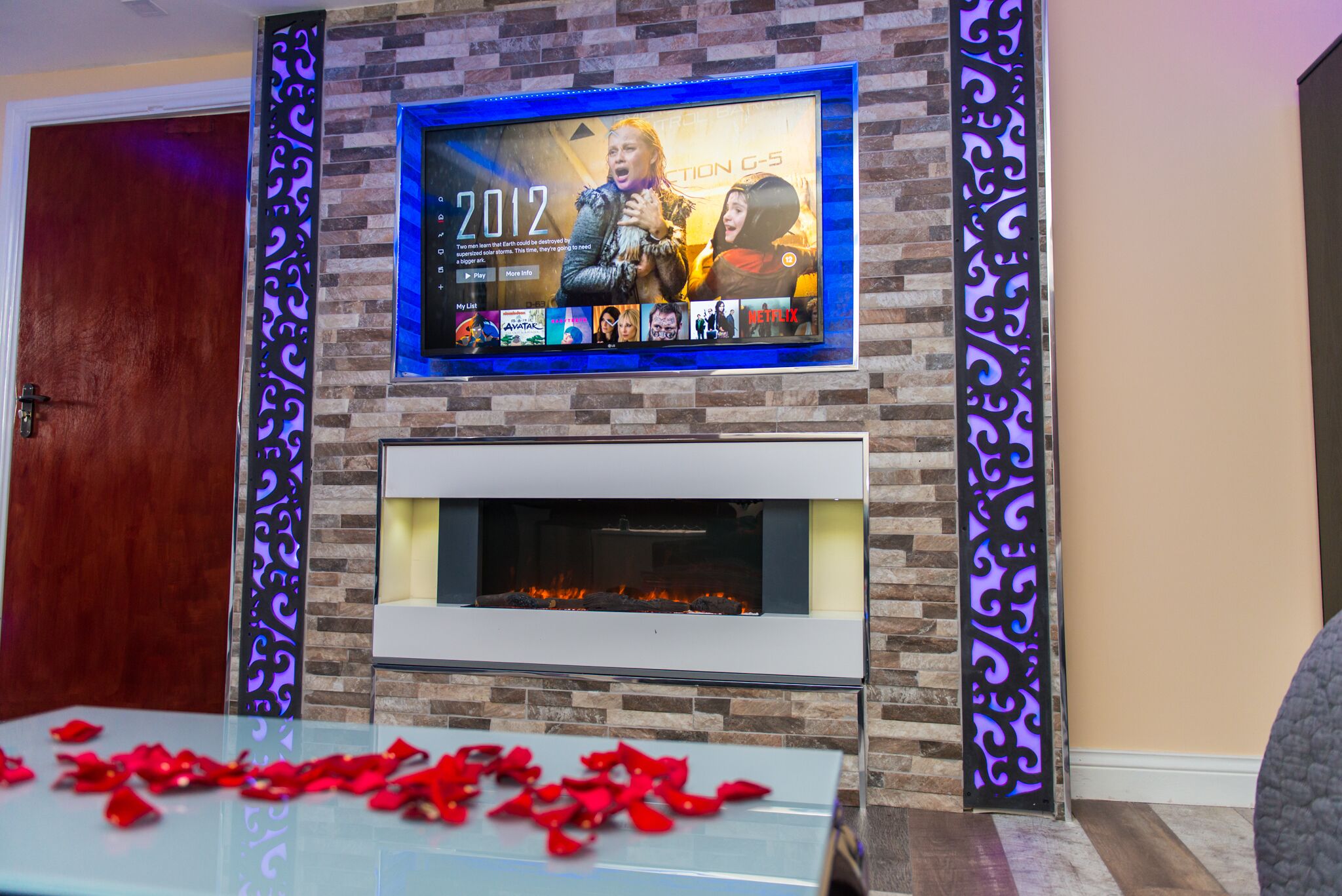 A living room with a fireplace, a TV mounted above it displaying the year 2012, and a glass table with rose petals.