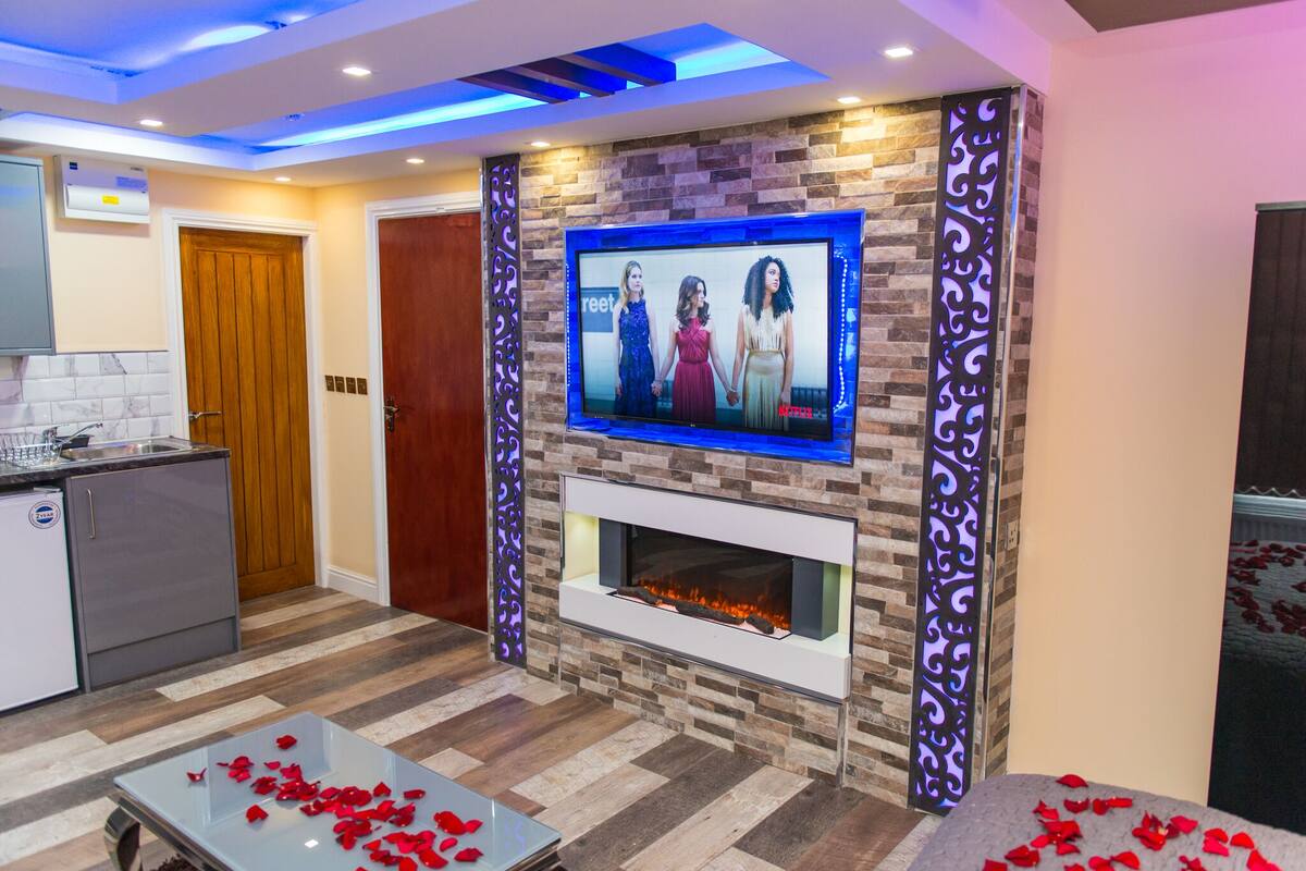 Una sala de estar moderna con chimenea, un televisor montado en la pared y una cocina visible a la izquierda.