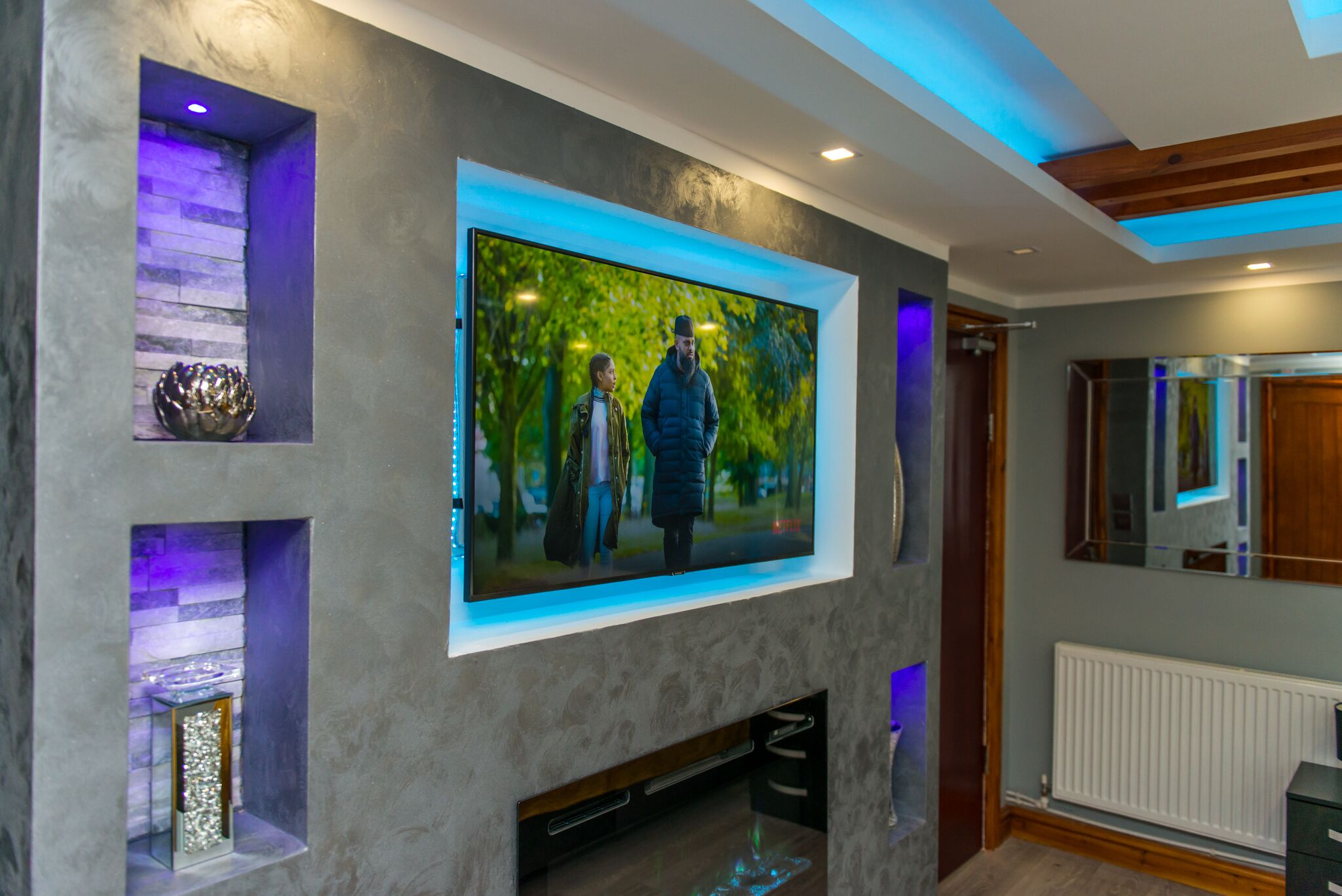 Ein modernes Wohnzimmer mit einem großen Flachbildfernseher an einer strukturierten Wand, einem Kamin darunter und eingebauten Regalen mit dekorativen Gegenständen.