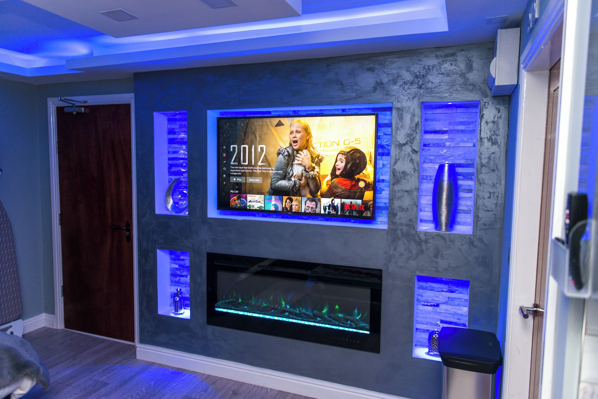 A modern living room with a large flat-screen TV mounted on the wall, a fireplace below, and a door to the left.