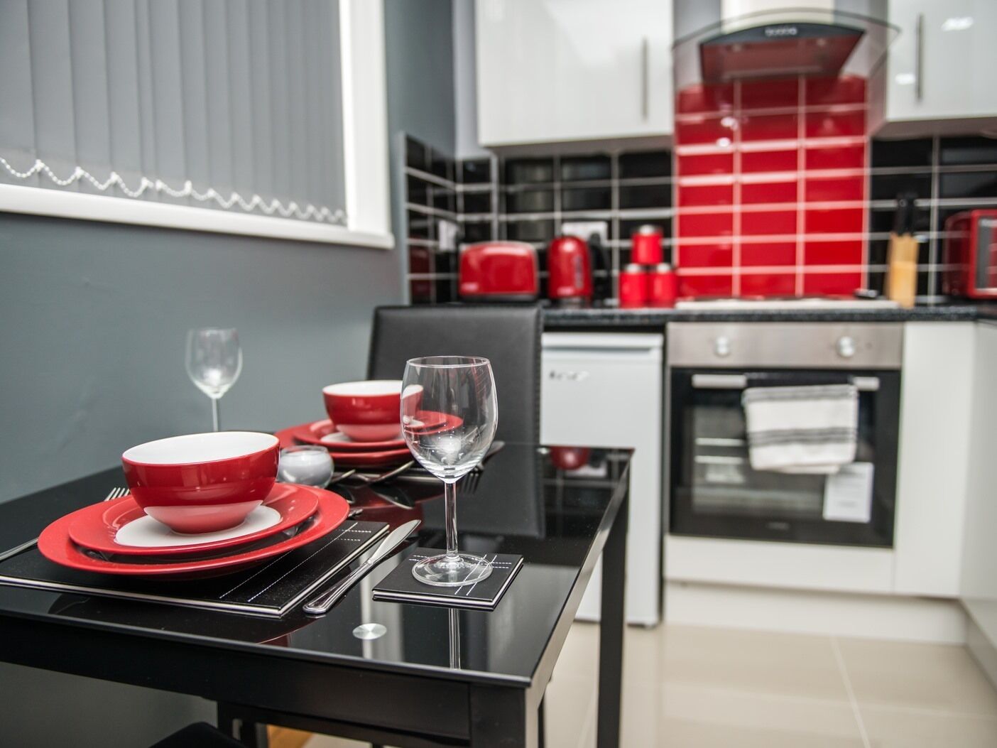A modern kitchen with red and white appliances, a black dining table set with red plates and a wine glass, and a view of the kitchen area.