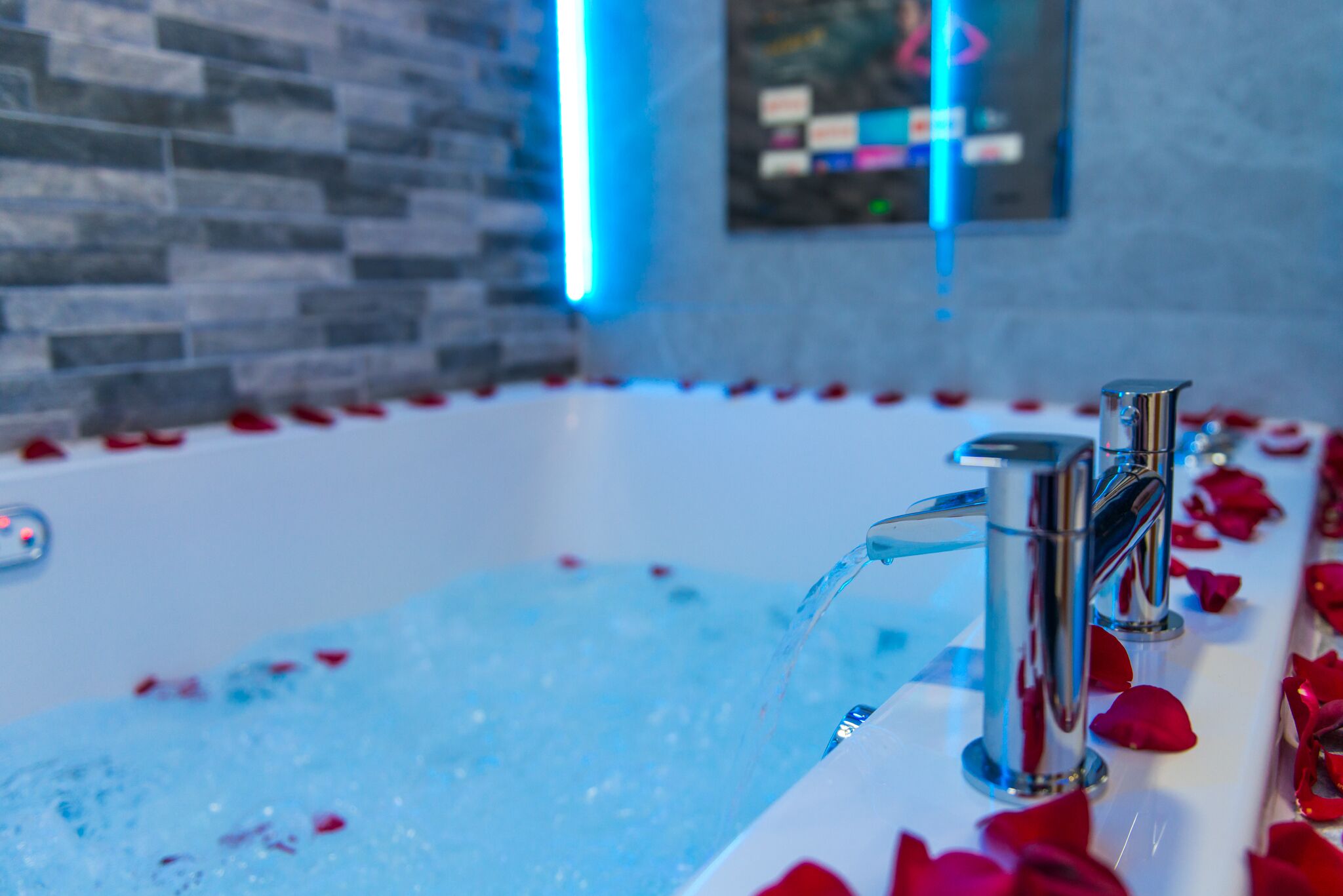 A bathtub filled with water and rose petals, with a stone wall and a framed picture in the background.