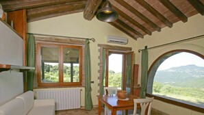 A cozy room with a white sofa, a wooden dining table with a chair, a view of the outdoors through a large window, and a ceiling with exposed wooden beams. - Beautiful private villa for 2 people with WIFI, A/C, TV, patio, pets allowed and panoramic view (Roccastrada)