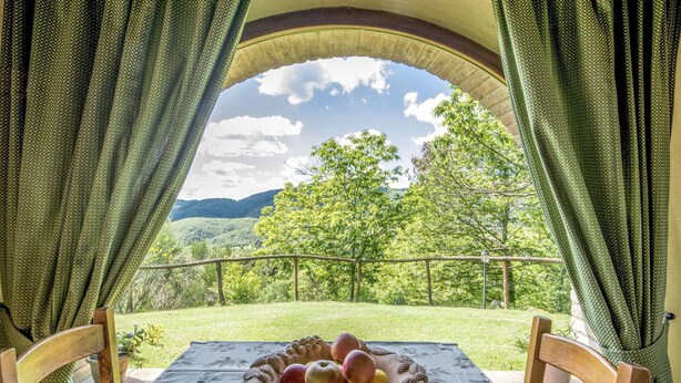 A table with a floral tablecloth and a fruit bowl, set in a room with a view of a green landscape and mountains through an arched window. - Beautiful private villa for 2 people with WIFI, A/C, TV, patio, pets allowed and panoramic view (Roccastrada)