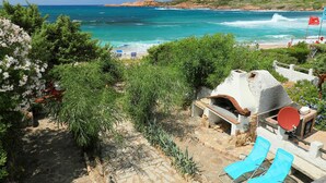 A stone pathway leading to a rustic outdoor seating area with a pizza oven, overlooking a beach with clear blue waters and rocky cliffs. - Cosy apartment for 3 guests with patio and pets allowed (Trinità D'agultu)