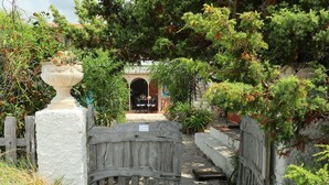 A rustic gate leading to a covered outdoor seating area with a table and chairs, surrounded by lush greenery and a stone wall. - Cosy apartment for 3 guests with patio and pets allowed (Trinità D'agultu)