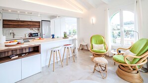 A modern kitchen with white cabinets, a wooden countertop, and a ceiling fan. There are two green chairs and a small stool in the living area.