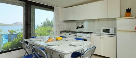 A compact kitchen with white cabinets, a dining table with blue chairs, and a view of the sea through a large window.