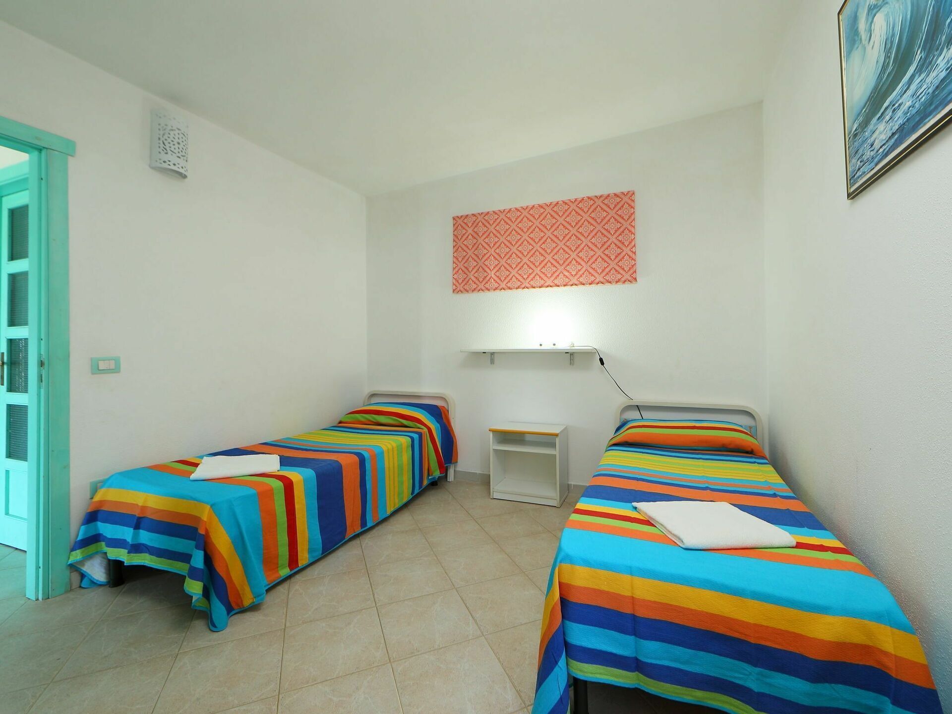 Ein Zimmer mit zwei Betten, jedes mit einer farbenfrohen, gestreiften Tagesdecke. Es gibt einen kleinen Schreibtisch mit einer Lampe, ein Wandbehang mit Muster und eine Tür mit türkisem Rahmen.
