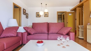 Una sala de estar con un sofá rojo, una mesa de centro blanca con un cuenco de fruta y copas de vino, y una lámpara.