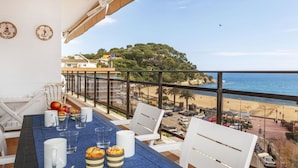 Un balcón con una mesa para dos, con vista a una playa y una colina boscosa.