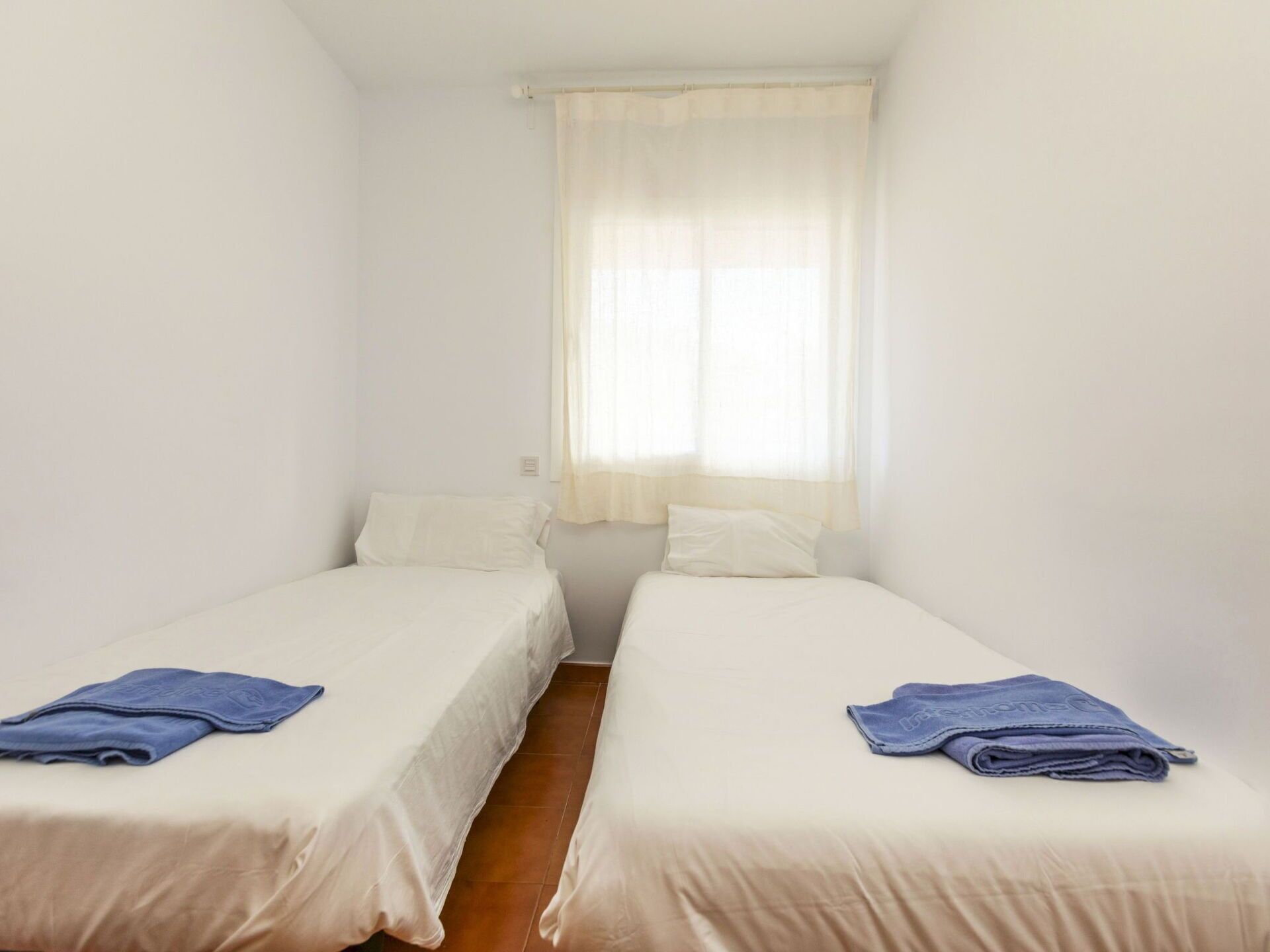 Ein kleines, sauberes Zimmer mit zwei Einzelbetten, einem Fenster mit durchscheinenden Vorhängen und einer schlichten weißen Wand.