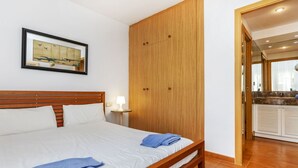 Una cama bien hecha con s ábanas blancas y toallas azules, un armario de madera, una mesita de noche pequeña con una lámpara y un cuadro en la pared.