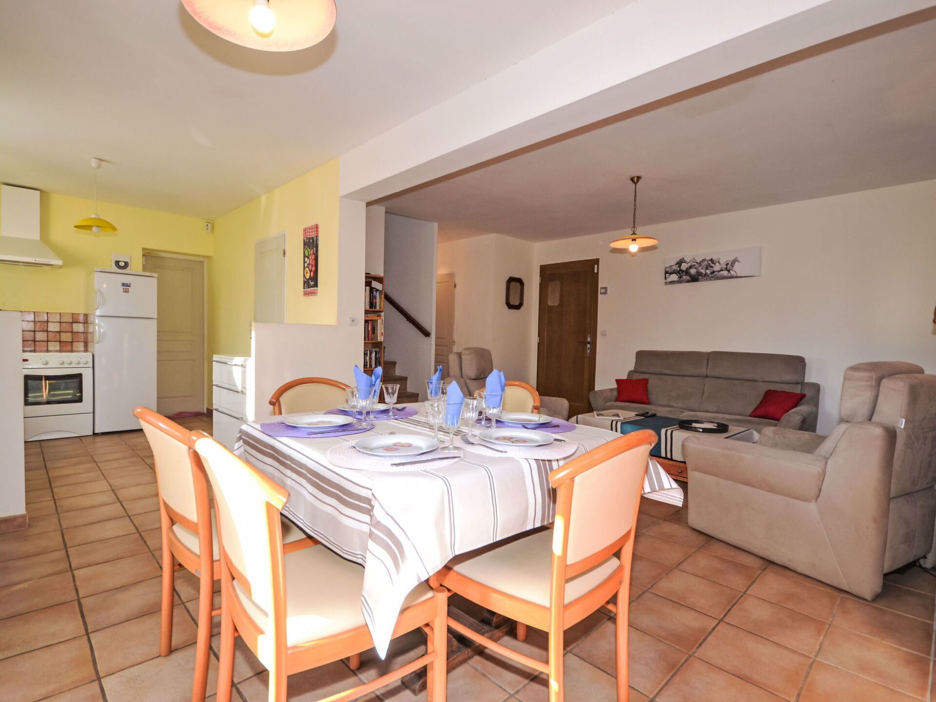A dining area with a table set for four, a kitchen with a refrigerator and stove, a sofa, and a staircase leading to another level.