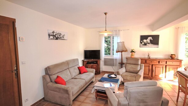 A living room with a sofa, armchairs, a coffee table, a TV, and a dining area with a cabinet and a lamp.