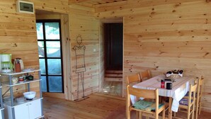 A wooden cabin interior with a dining table, chairs, a kitchen area with shelves, and a large window. - Kerca Bio Farm (Kercaszomor)