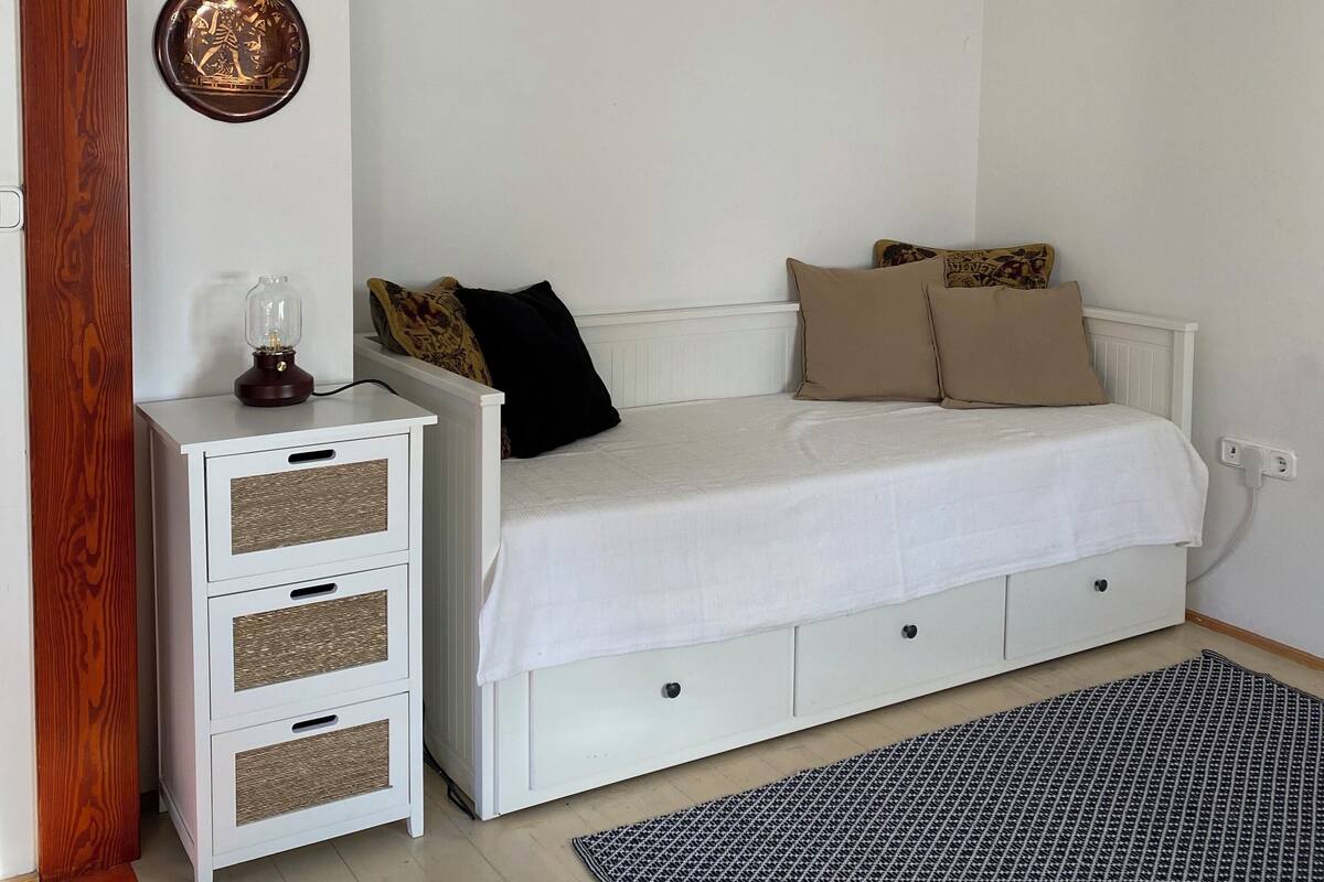 A bedroom with a white bed, a nightstand with drawers, a wall-mounted light, and a patterned rug.
