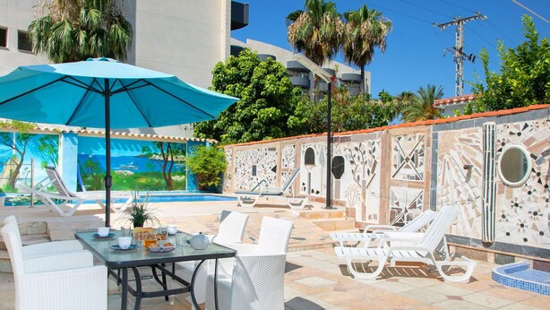 Ein Poolbereich mit Liegestühlen, einem Tisch mit blauem Sonnenschirm und einer Wand mit Mosaikfliesen.