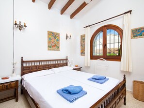 A double bed with white linens, two blue towels, a wooden headboard, a nightstand with a lamp, a wall-mounted light fixture, a window with a wooden frame, and two decorative wall hangings. - Wonderful private villa for 4 guests with private pool, A/C, WIFI, TV, terrace and panoramic view (Teulada)