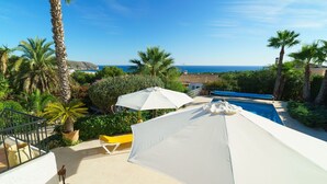 Un espace piscine avec vue sur la mer, entouré de palmiers et agrémenté d’un parasol blanc.
