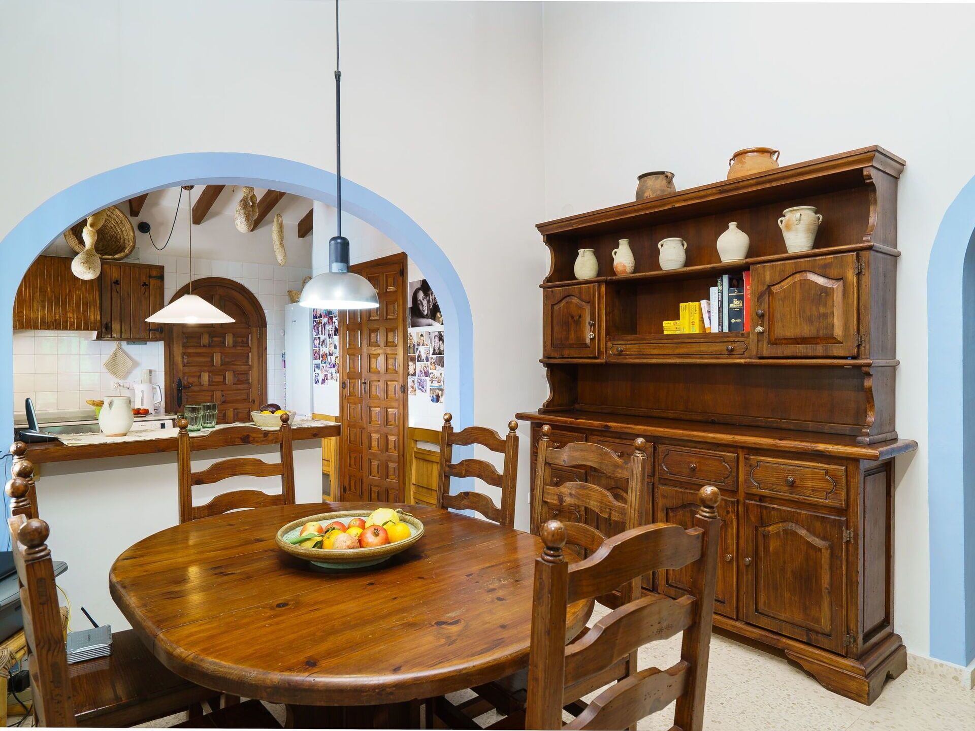 Een eethoek met een houten tafel, stoelen en een schaal met fruit. Daarboven staat een houten kast met planken.