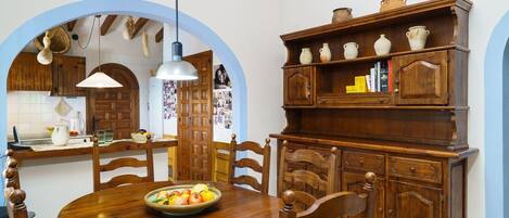 Een eethoek met een houten tafel, stoelen en een schaal met fruit. Daarboven staat een houten kast met planken.