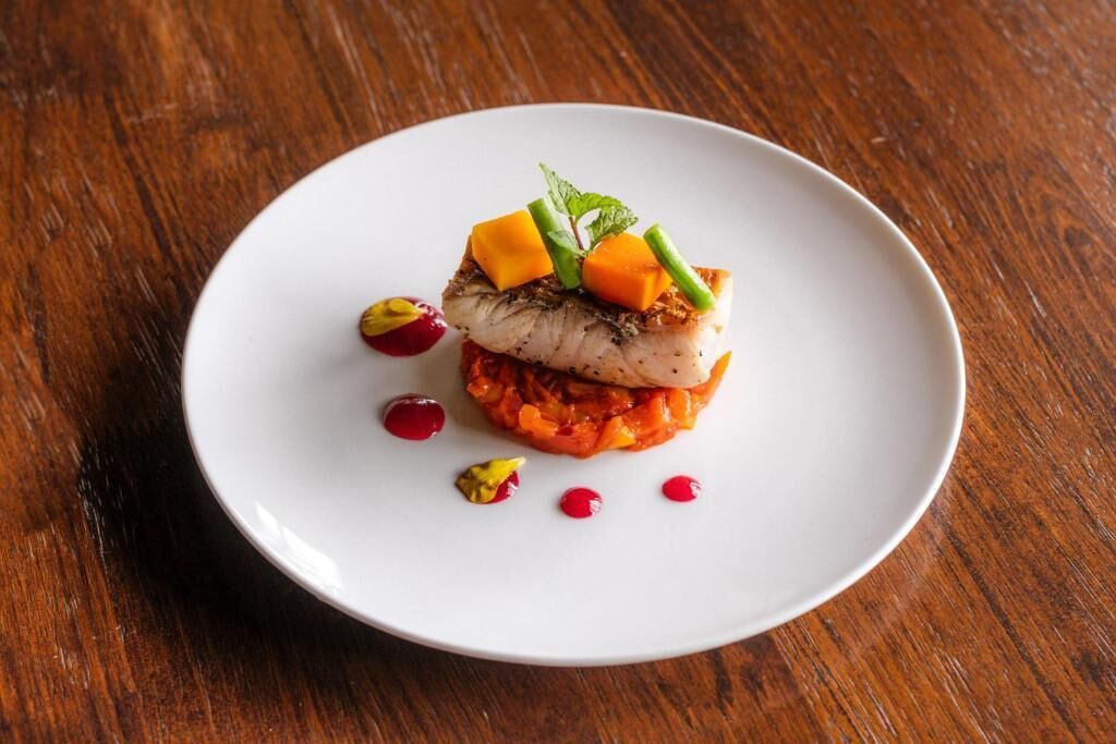 A white plate with a piece of fish topped with mango and green beans, garnished with mint, on a wooden table.