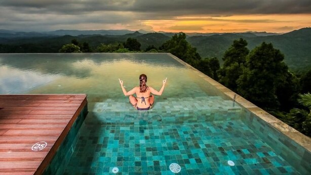 A person sitting in a clear-bottomed infinity pool with a wooden deck on one side, surrounded by lush greenery and mountains in the background. - Simpson's Forest Hotel (Wattegama)