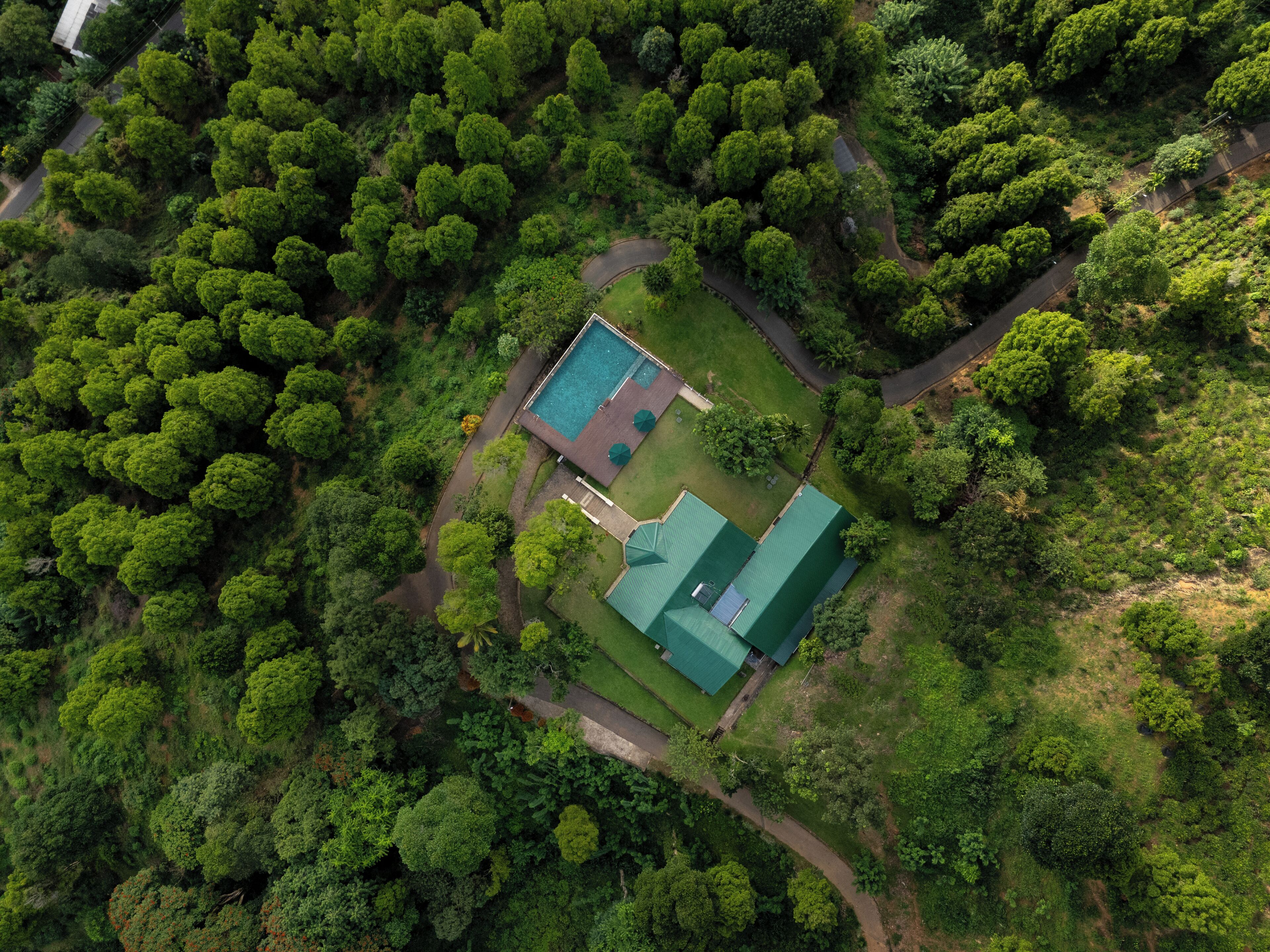 An aerial view of a property surrounded by dense greenery, featuring a swimming pool, a tennis court, and a large building with a green roof.