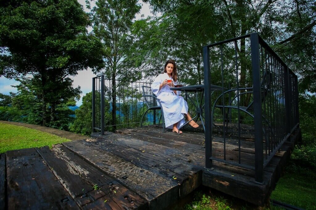 A person sitting on a wooden platform with a metal railing, surrounded by greenery and trees.
