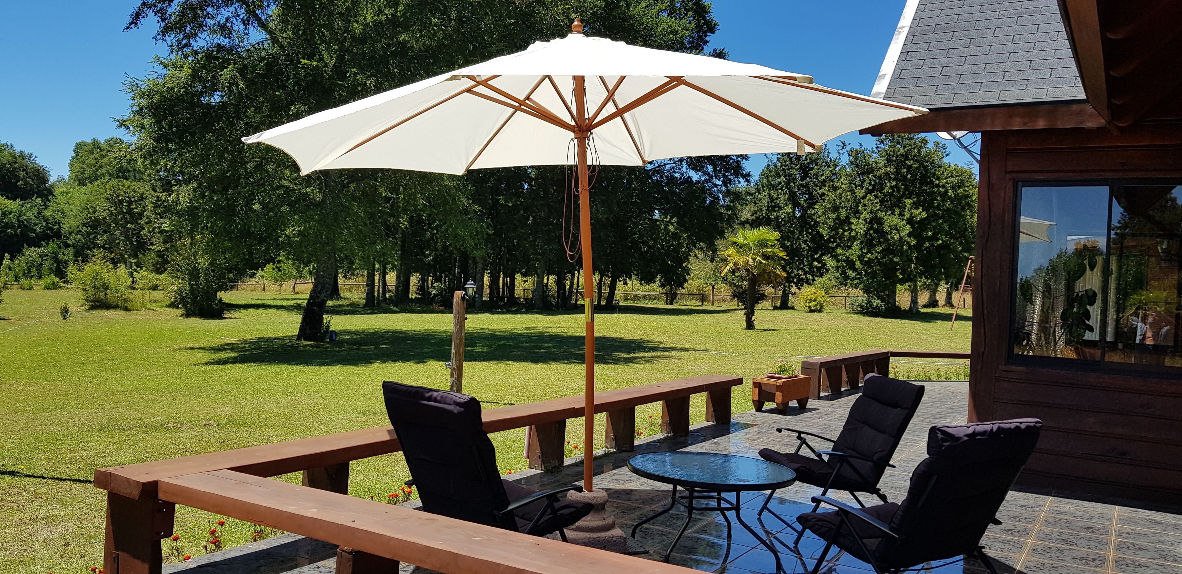 Un patio al aire libre con bancos de madera, una mesa redonda y un paraguas grande.