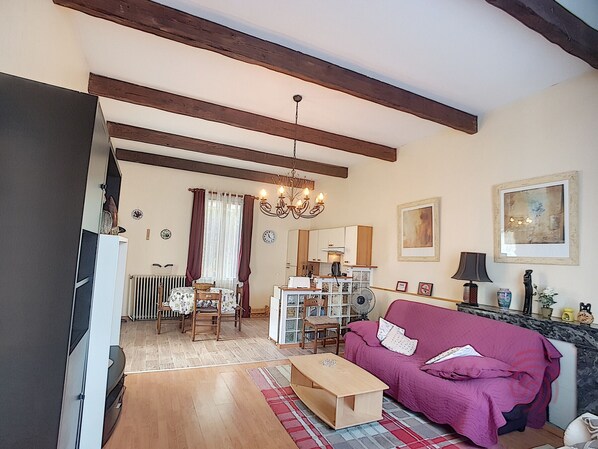 A living room with a wooden ceiling, a chandelier, a purple sofa, a wooden coffee table, and a dining area with a table and chairs.