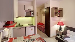 Habitación de hotel compacta con kitchenette, una cama, una mesita de noche con lámpara y un espejo pequeño.
