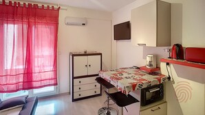 Una habitación pequeña y bien iluminada con una cama, un escritorio, una silla, un televisor y una kitchenette.