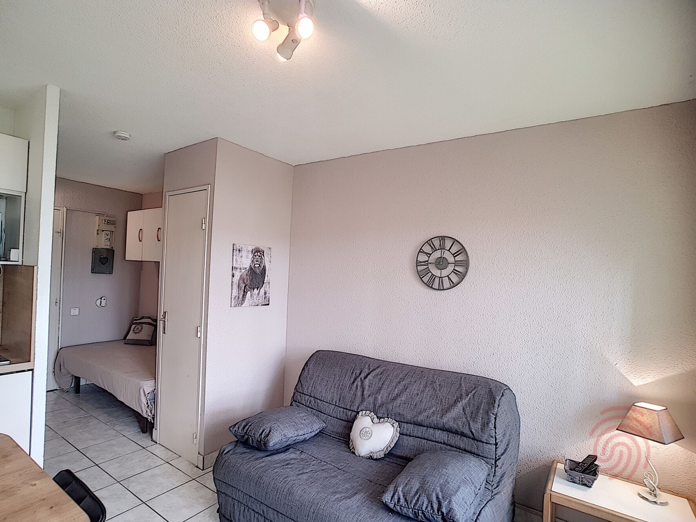 Un dormitorio con un sofá gris, un reloj de pared, un cuadro de un león y una mesita de noche con una lámpara.