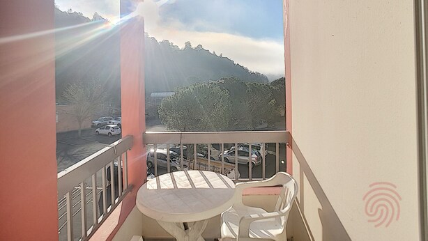 Un balcon avec une table et des chaises, d’où l’on peut admirer un parking et un paysage vallonné.