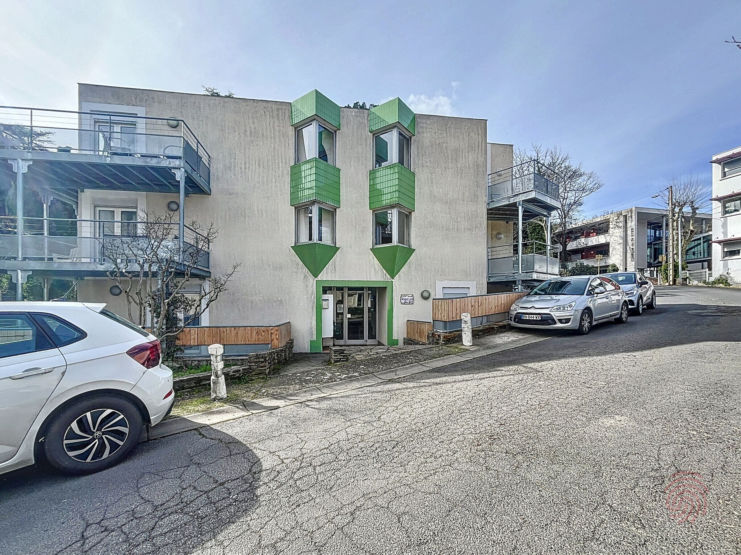 Ein Gebäude mit modernem Design, das ein flaches Dach und einen großen Eingangsbereich mit einem grünen Markisenvorbau aufweist. Vor dem Gebäude sind mehrere Autos geparkt, und es scheint Teil eines größeren Komplexes mit mehreren Gebäuden zu sein.