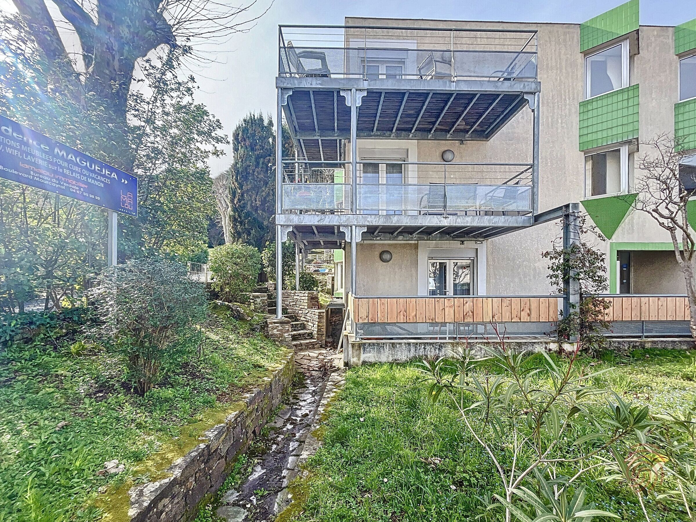 A two-story building with a balcony, surrounded by greenery and a small stream.